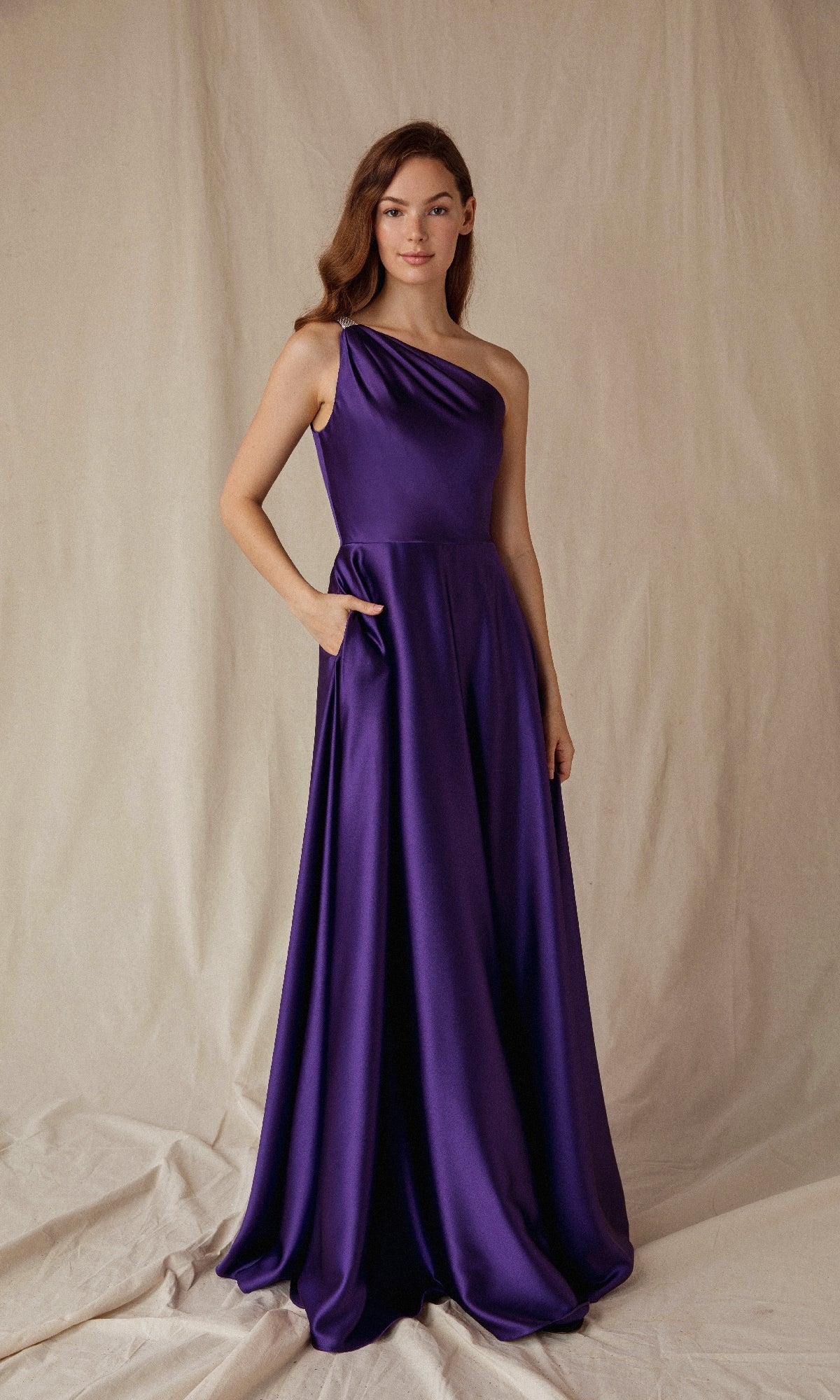 Standing against a neutral backdrop, a person wears the Eureka 9025—an elegant, floor-length one-shoulder satin formal dress in deep purple with pockets, perfect for prom or evening events.
